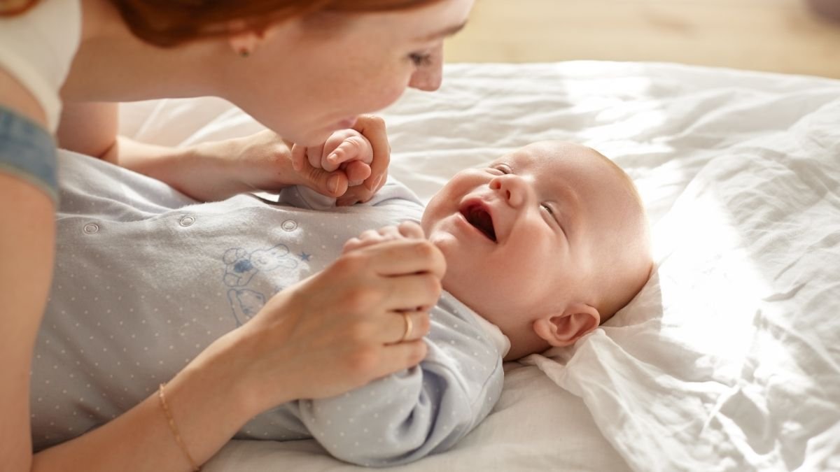 Mãe debruçada sobre a cama brincando com bebê de poucos meses que sorri