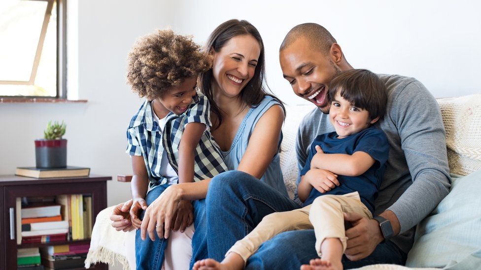 Pai e mãe sentados no sofá rindo com dois filhos pequenos no colo
