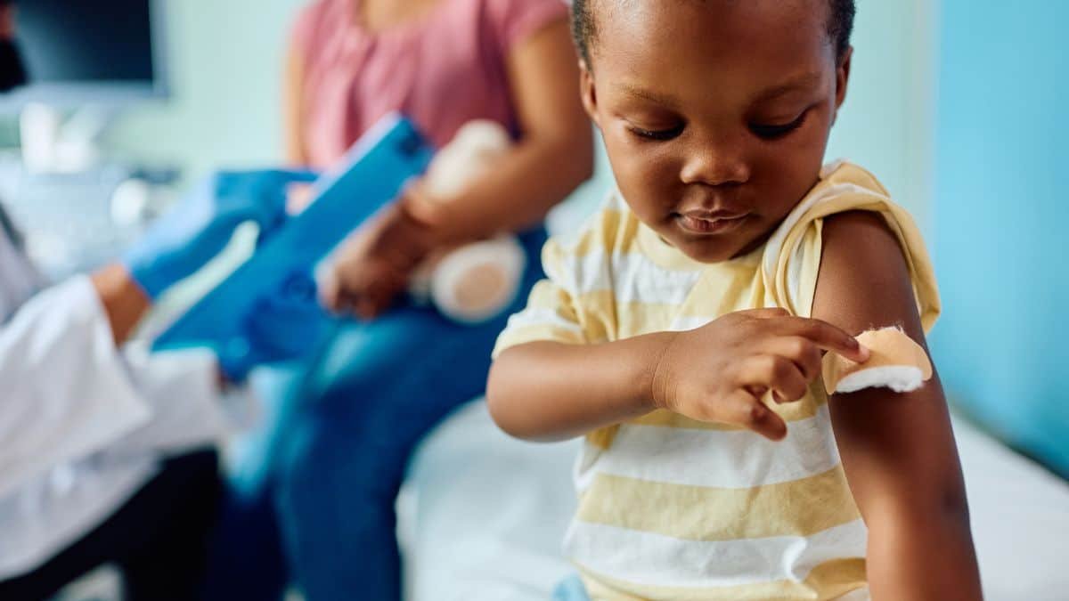 Criança pequena olhando o curativo no braço após tomar vacina, com profissional de saúde ao fundo